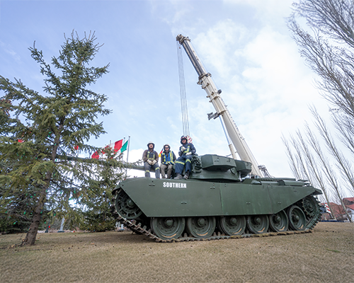 Three workers sitting on a green military tank named 'Southern' with a McIntyre crane in the background, surrounded by trees and flags